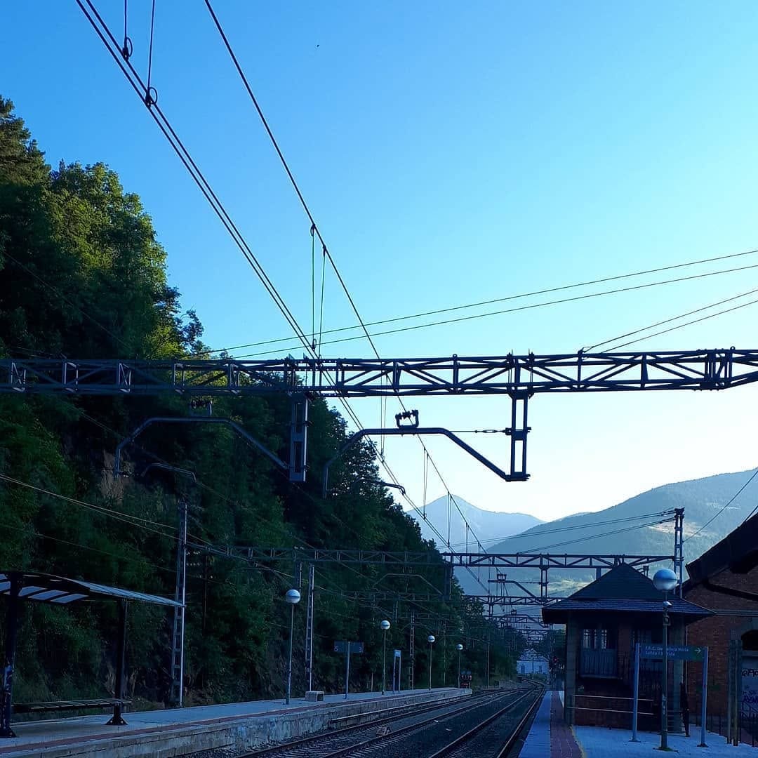 Paisaje de una estación de tren de montaña al atardecer: vías y andén en primer plano, catenaria y estructura metálica de la línea eléctrica cruzando el cielo celeste despejado, y una…