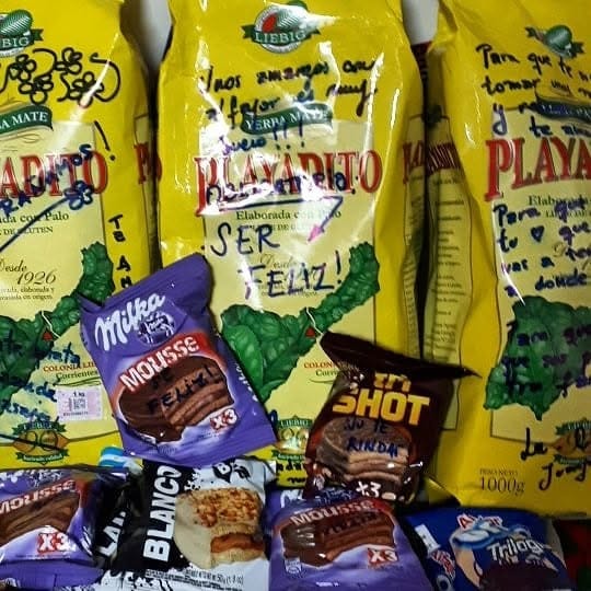 Bodegón cenital de paquetes de yerba mate Liebig "Playadito" y golosinas (Milka Mousse, Tri Shot, Trilogy), intervenidos a mano con dibujos y frases en marcador negro y rosa, como "ser…
