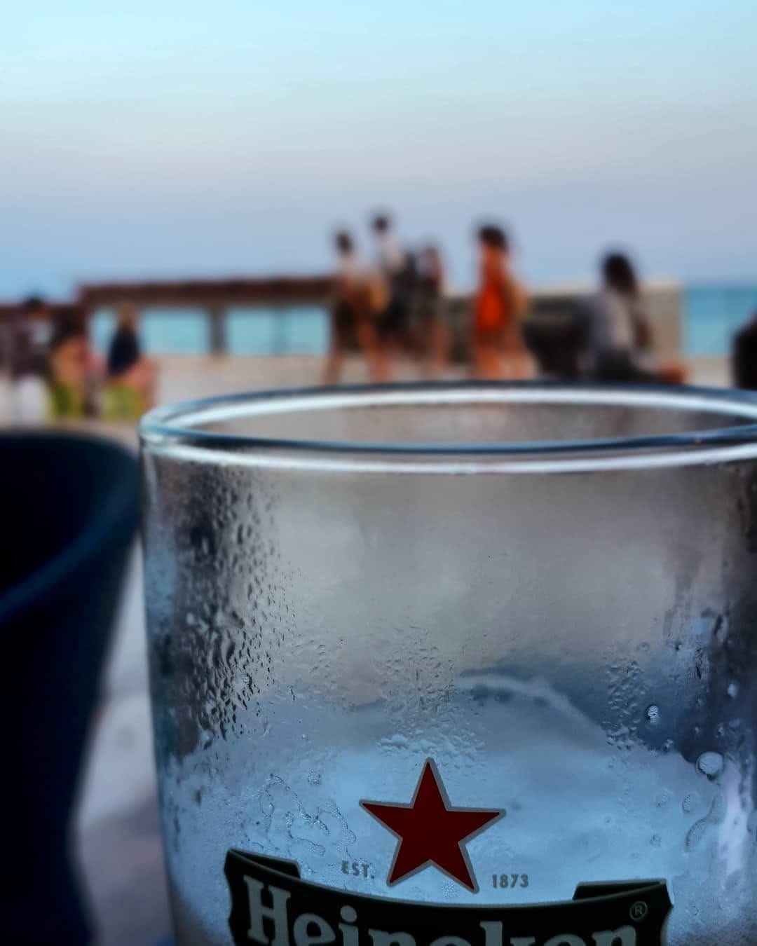 Primer plano de un vaso de cerveza Heineken empañado y con gotas de condensación, en primer término desenfocando un grupo de personas de pie junto a una baranda frente al mar al atardecer.