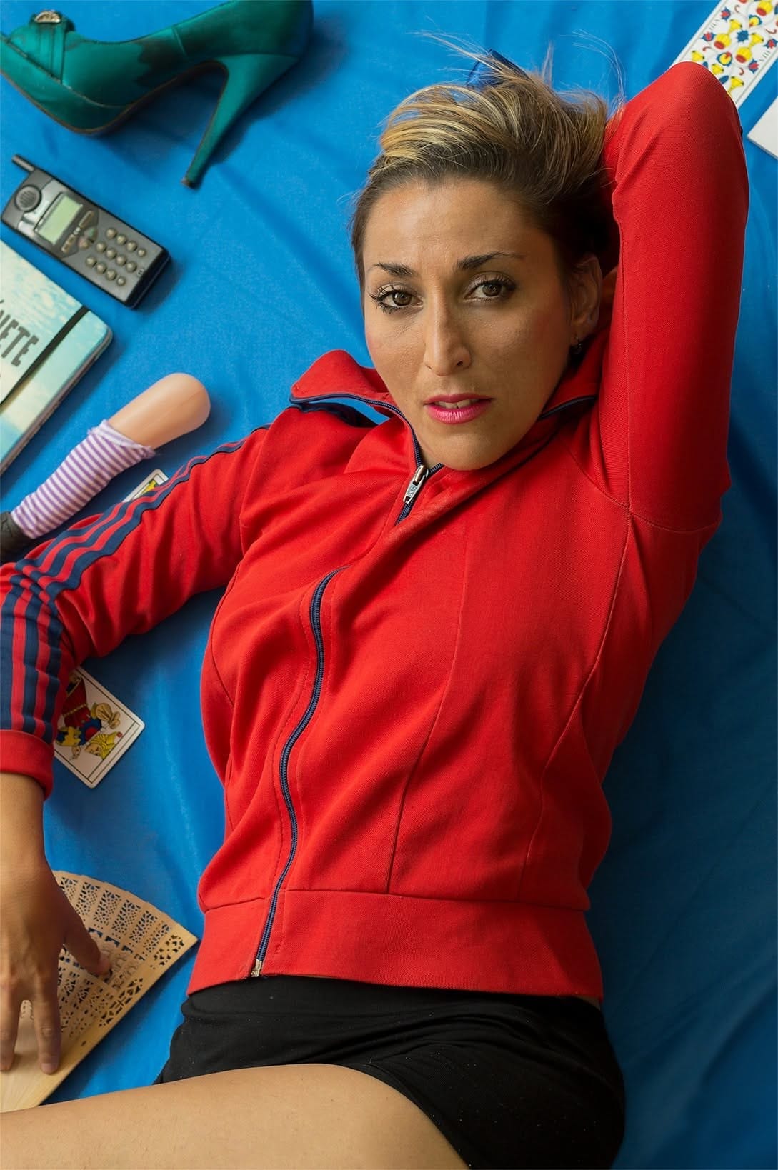 Retrato cenital de una mujer tendida sobre una manta azul, con campera roja de gimnasia con capucha, short negro y mirada firme a cámara.