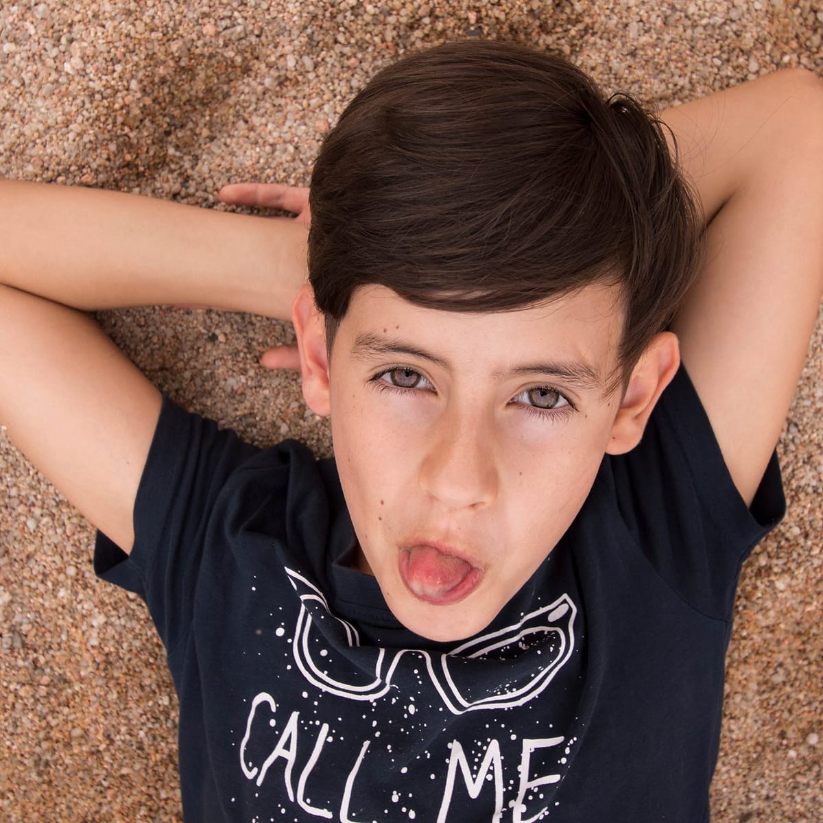Retrato cenital de un niño tumbado sobre arena de grano grueso, con las manos cruzadas bajo la nuca y la lengua afuera en gesto burlón.