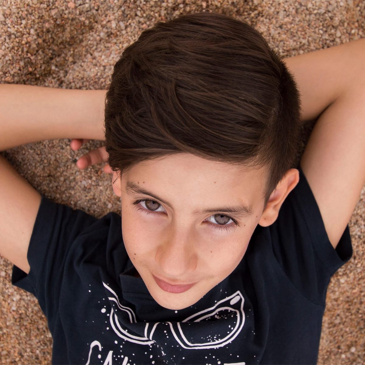 Retrato cenital de un adolescente tendido en la arena, con los brazos cruzados detrás de la cabeza y la mirada directa a cámara.