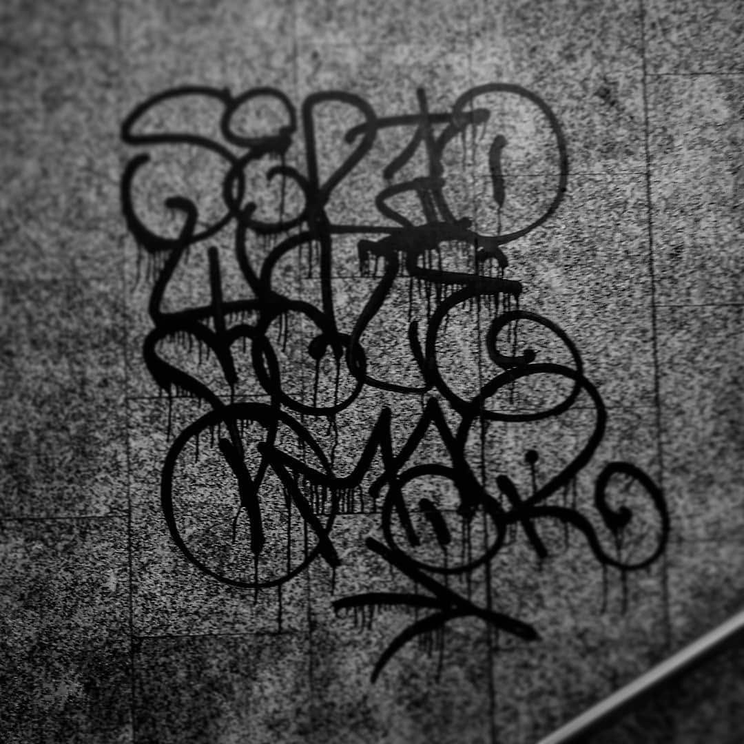 Detalle de un graffiti tipo tag en negro, con chorreados de pintura, sobre una pared de baldosas grises.