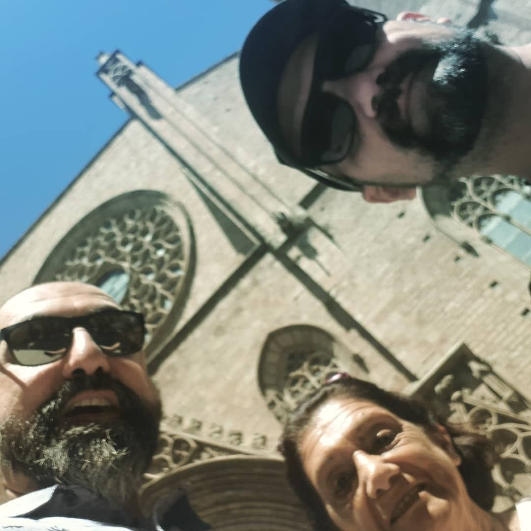 Selfie grupal en contrapicado extremo de tres personas sonrientes, con una fachada gótica de piedra y un gran rosetón de fondo bajo un cielo azul intenso.