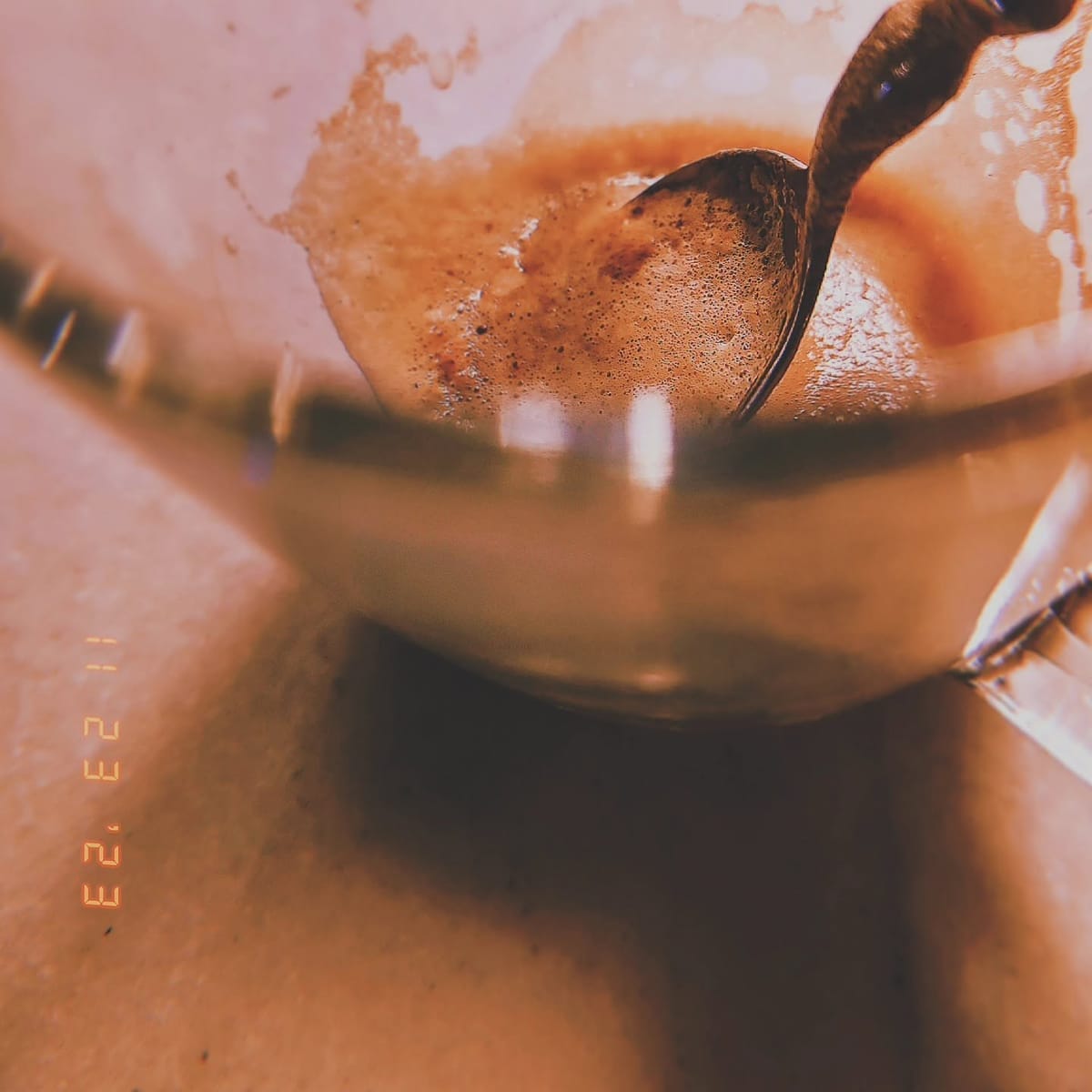 Macrofotografía en tonos cálidos ámbar de una taza de café con espuma, vista desde un ángulo bajo con una cuchara metálica apoyada en el borde.