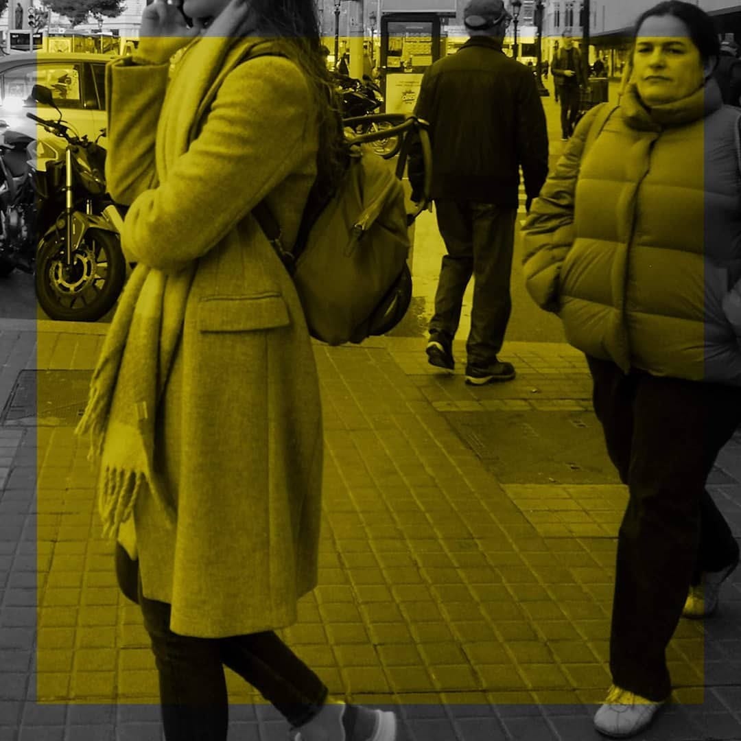 Escena callejera en blanco y negro con viraje amarillo, donde tres personas caminan en distintas direcciones por una vereda: una mujer con abrigo y mochila hablando por telefono en primer…