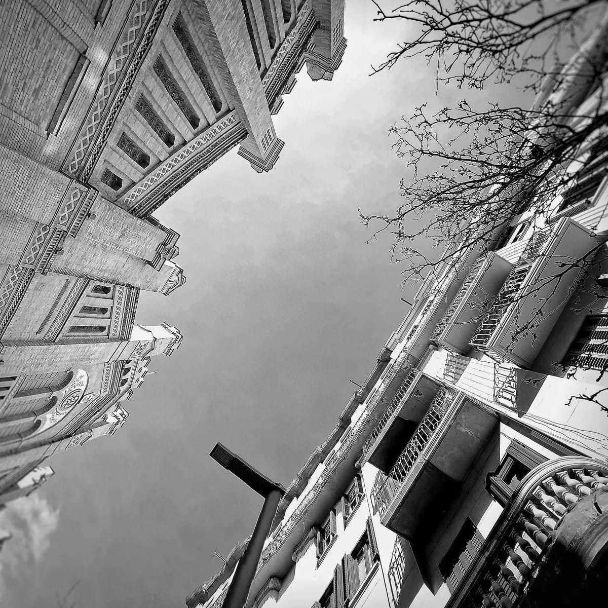 Fotografia en blanco y negro tomada en gran angular mirando hacia arriba, donde varias fachadas de edificios historicos con balcones y molduras convergen hacia un cielo gris cubierto, con…