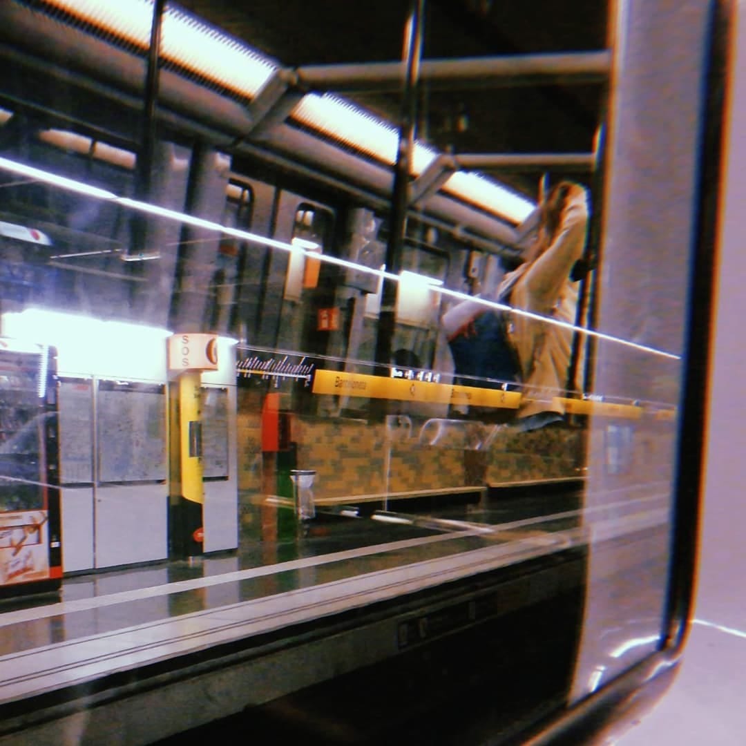 Fotografia tomada a traves de una ventanilla de metro que refleja el interior del vagon superpuesto sobre el anden de la estacion, donde se lee el cartel amarillo 'Barceloneta'.