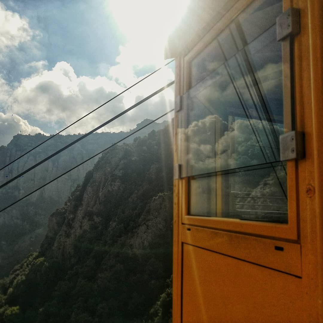 Interior de una cabina de teleferico de madera con vidrio, desde donde se ve un paisaje de montanas rocosas y vegetacion bajo un cielo con grandes nubes y fuerte contraluz solar.