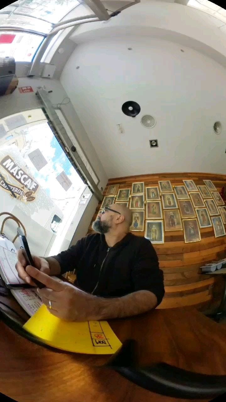 Toma con lente ojo de pez dentro de un local con techo curvo blanco y una pared cubierta de retratos enmarcados en tono sepia; un hombre calvo con barba y gafas, vestido de negro, escribe…