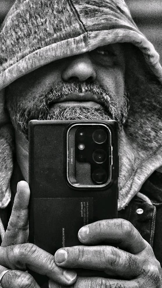 Primero un autorretrato en blanco y negro de alto contraste: un hombre con capucha sostiene un teléfono frente al rostro, apenas visible bajo la sombra de la capucha, con la marca de…
