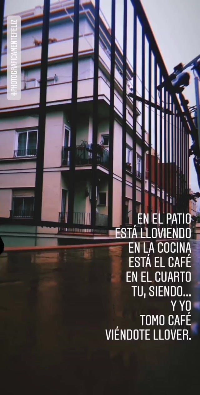 Plano fijo de una fachada de edificio de apartamentos vista a través de una reja o valla metálica en primer plano, con la calle mojada reflejando el cielo nublado.