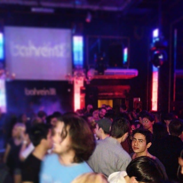 Interior de una discoteca abarrotada de gente, con una pantalla que muestra el logo "Bahrein" al fondo y luces de neón azules y rojas.