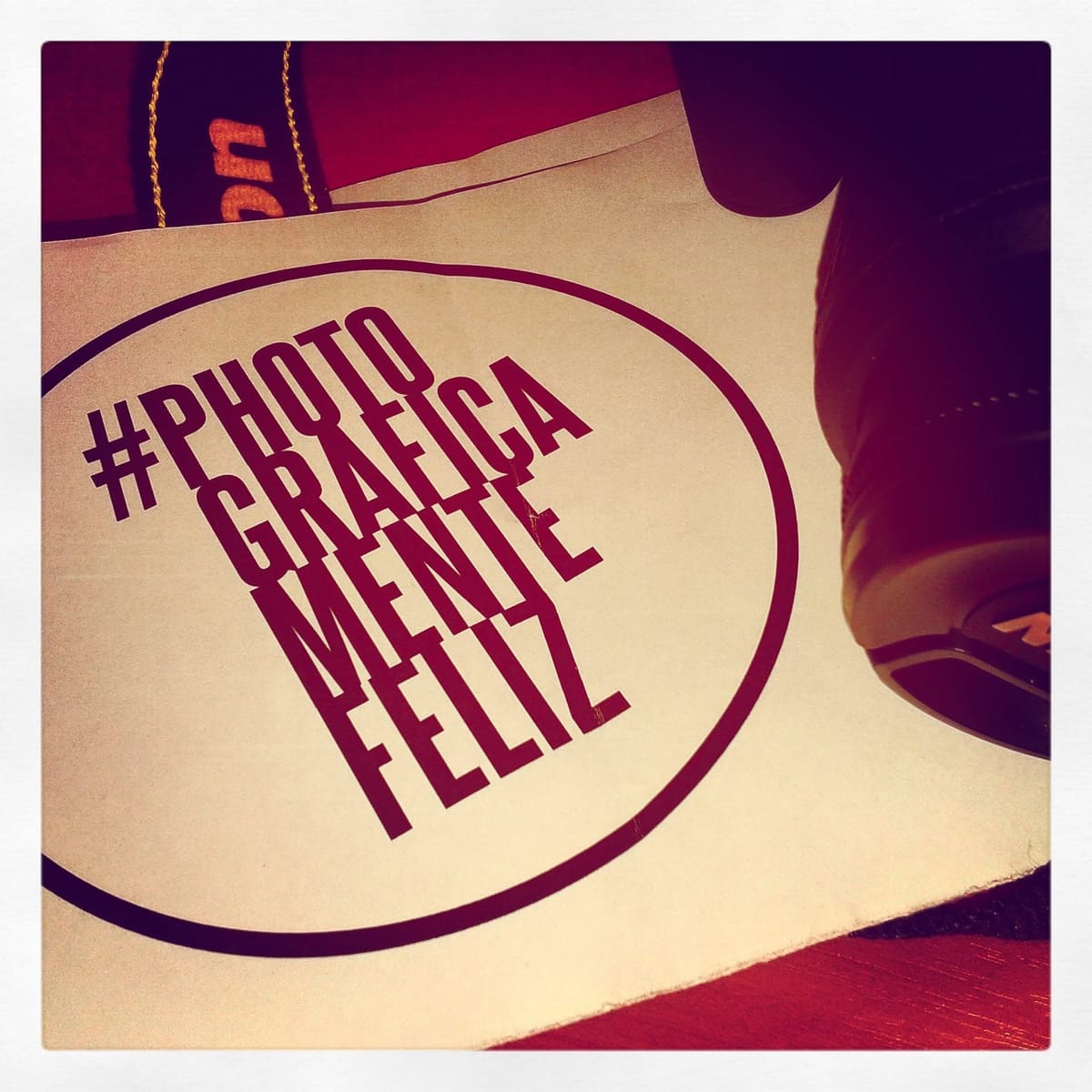 Fotografía de una tarjeta impresa con el logo "#photograficamentefeliz" en un óvalo bordó sobre papel claro, apoyada junto al borde de una zapatilla oscura sobre un piso rojizo.