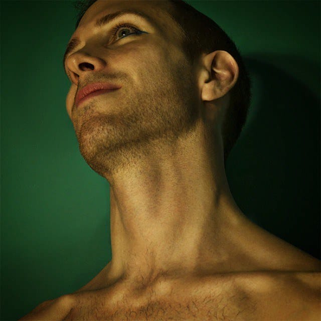Retrato de un hombre joven de perfil, mirando hacia arriba, con un fino delineado azul bajo los ojos y torso desnudo, sobre fondo verde oscuro liso.