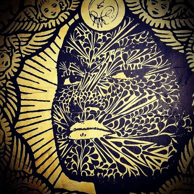 Mural de arte callejero en negro y dorado que representa un rostro estilizado rodeado de motivos florales y rayos, enmarcado por dos figuras de ángeles pequeños en las esquinas superiores.