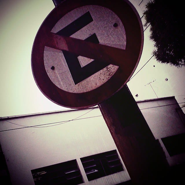 Vista en contrapicado de un cartel vial circular de "prohibido estacionar" oxidado, montado en un poste de madera oscura, con un edificio bajo y un árbol al fondo bajo cielo claro.