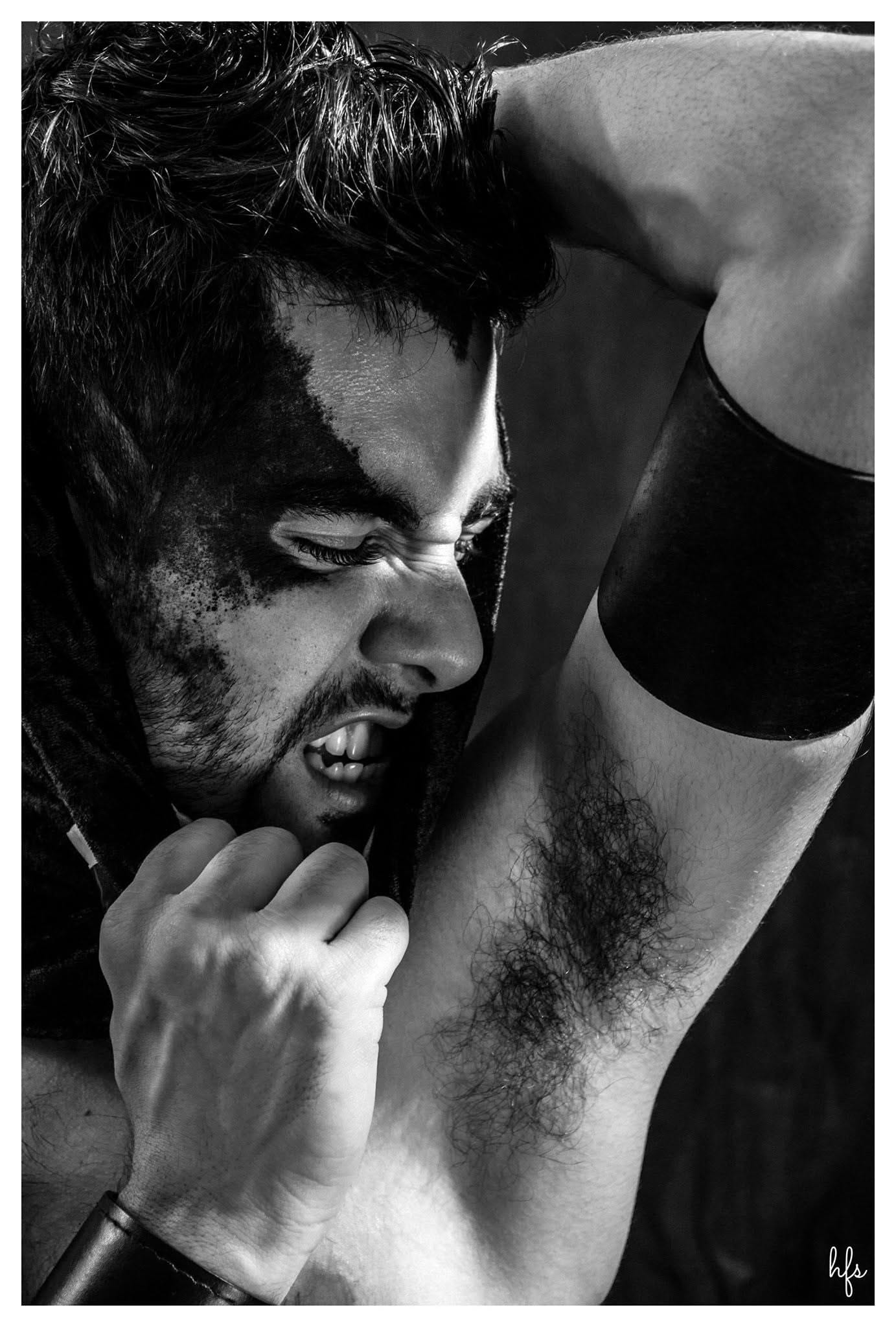 Retrato dramático en blanco y negro de un hombre con el rostro parcialmente sucio y en sombra, gesto de tensión mordiéndose los nudillos, apoyado contra su propio brazo levantado con una…