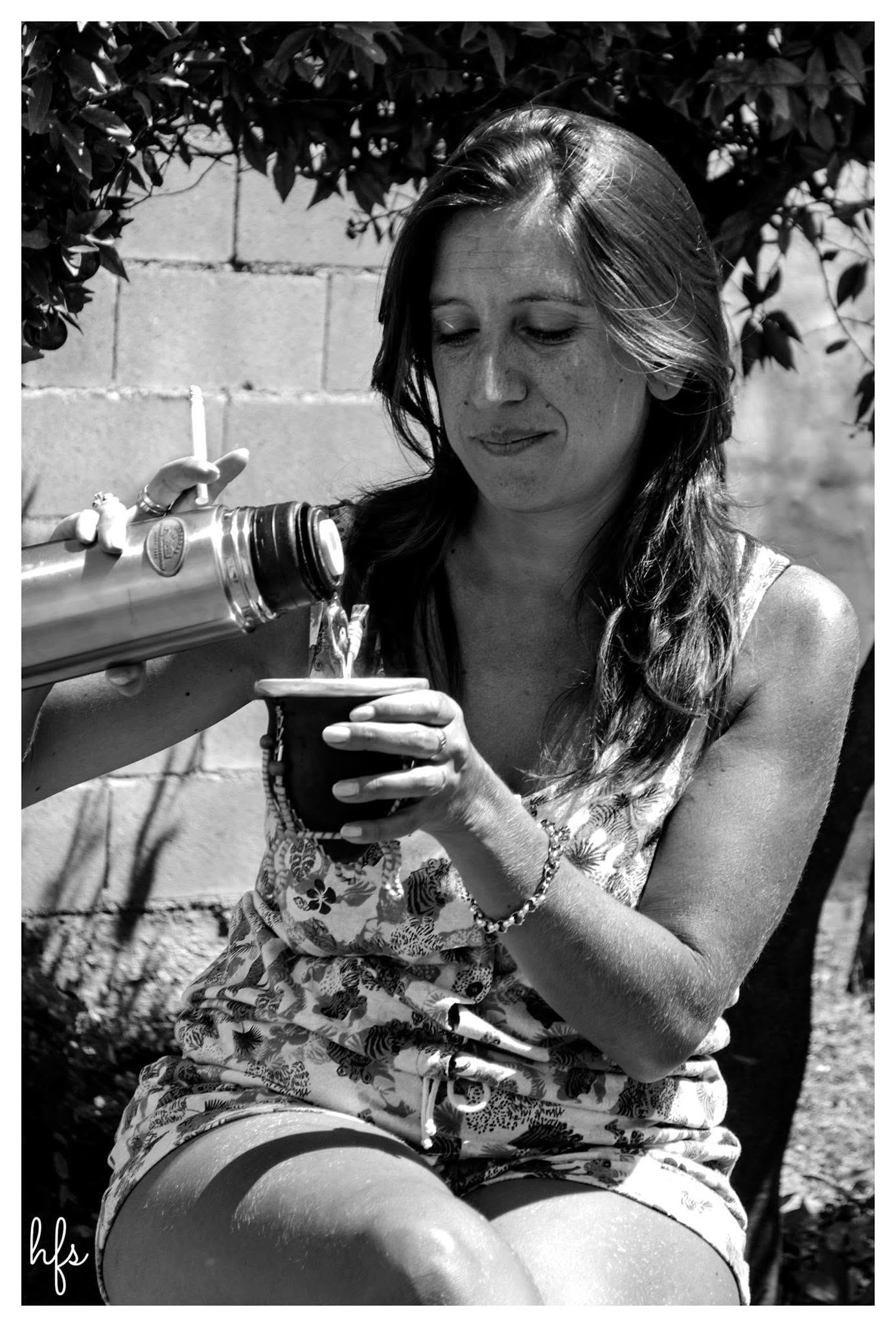 Fotografía en blanco y negro de una mujer de pelo oscuro sentada bajo un árbol, sirviéndose agua de un termo en un mate; viste un vestido sin mangas floreado y pulsera, con una pared de…