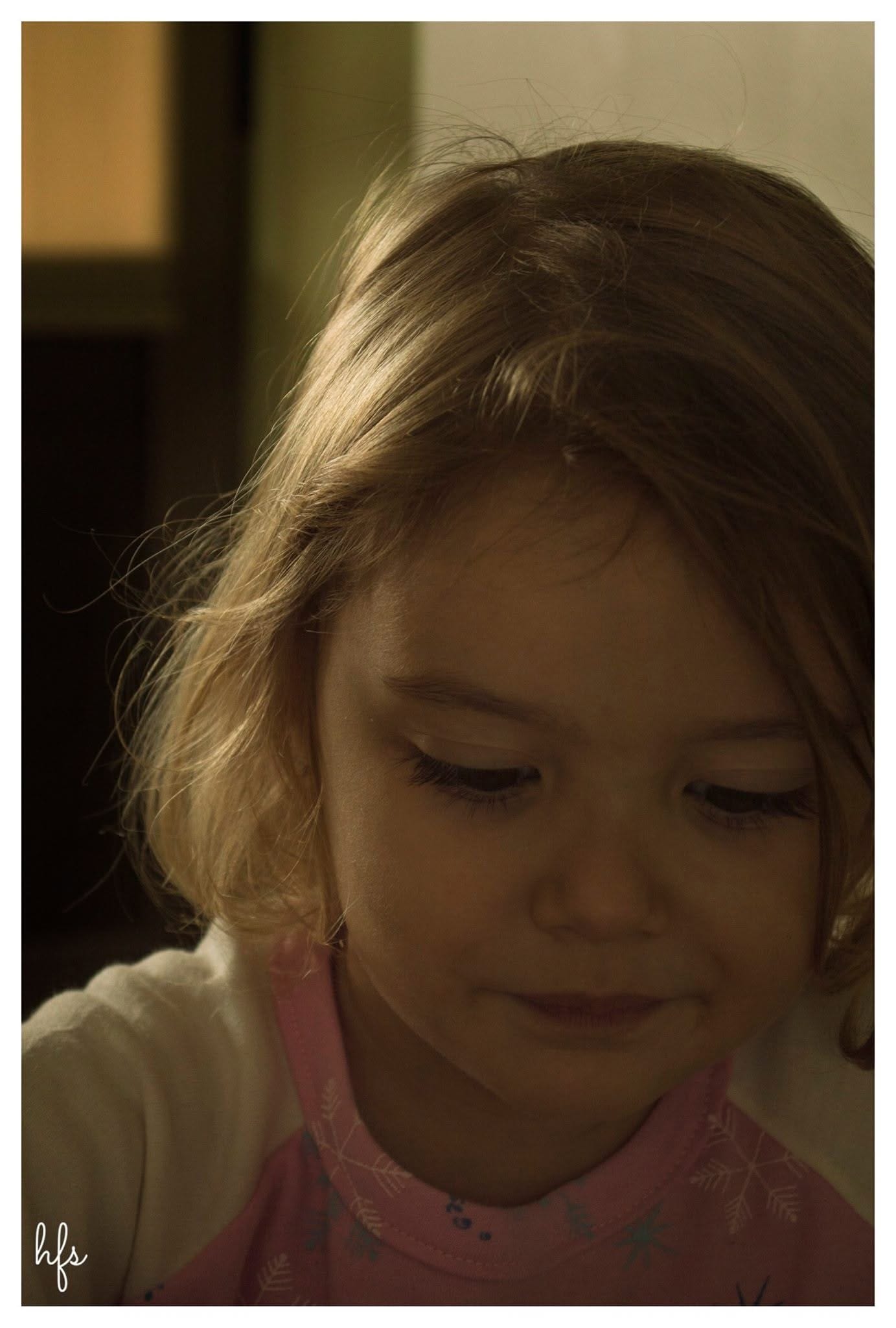 Retrato a color en primer plano de una niña pequeña de pelo castaño claro iluminado por luz cálida lateral, mirando hacia abajo con una leve sonrisa; viste una prenda rosa con estampado de…