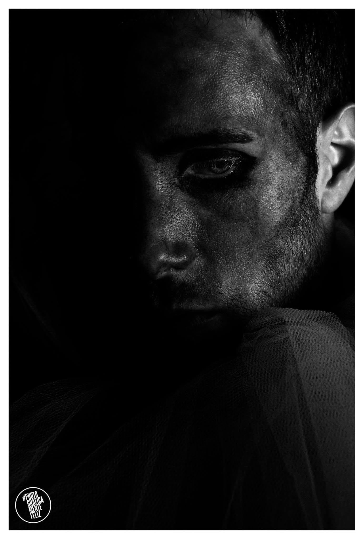 Retrato de un hombre sonriente con la cabeza cubierta por un velo o tul verde translúcido, con maquillaje oscuro corrido bajo los ojos y barba, mirando hacia un costado con expresión cálida.