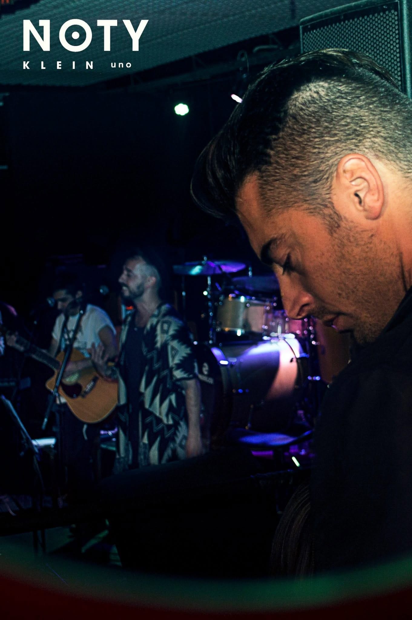 Retrato de perfil de un joven de pelo peinado hacia atrás, mirando hacia abajo, con un músico tocando guitarra y batería desenfocados al fondo bajo luces de escenario.