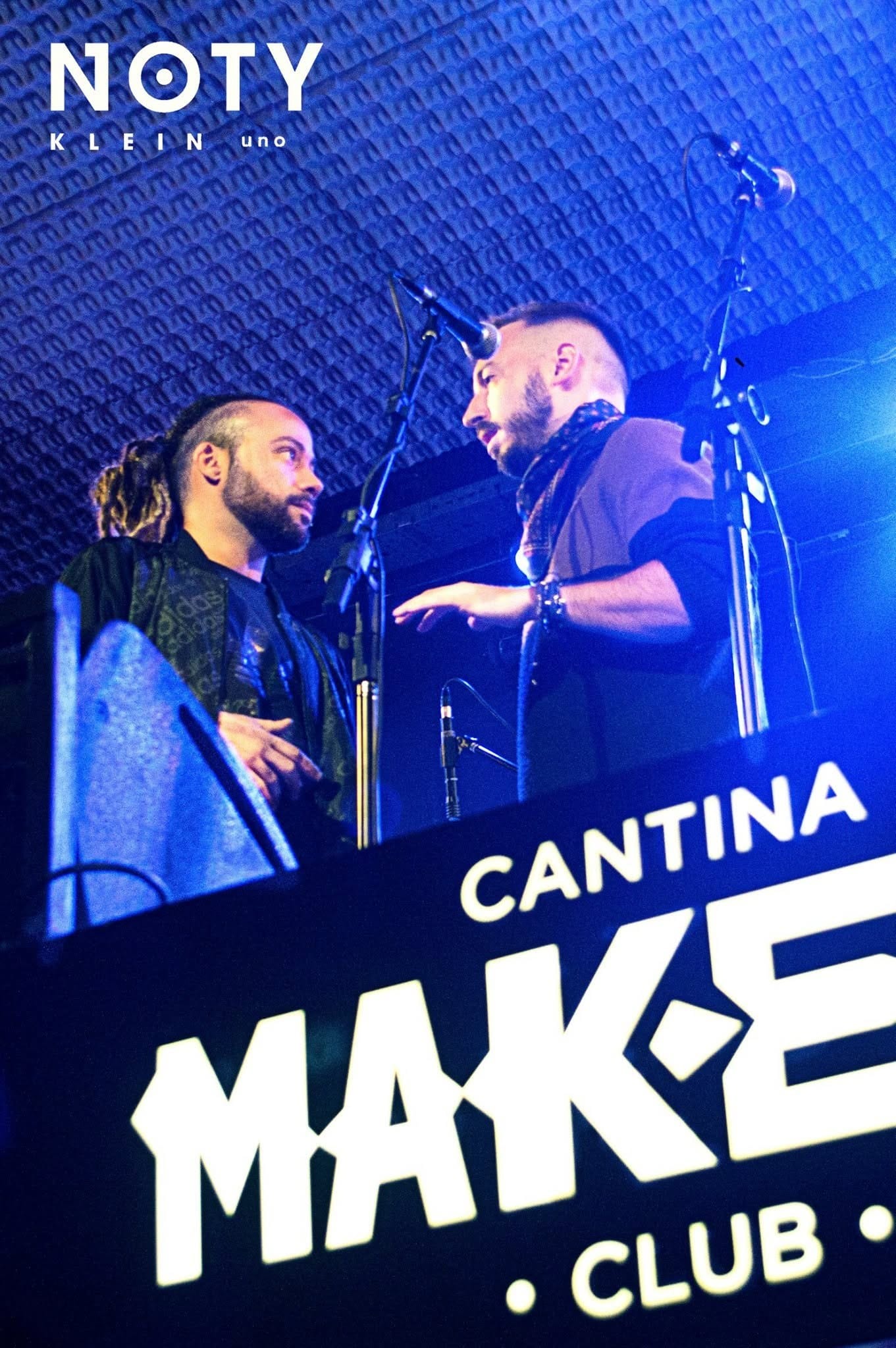 Fotografía a color de dos hombres conversando de cerca junto a sendos micrófonos sobre un escenario con fondo texturado azul, uno con corte de pelo rapado a los costados; abajo se lee…
