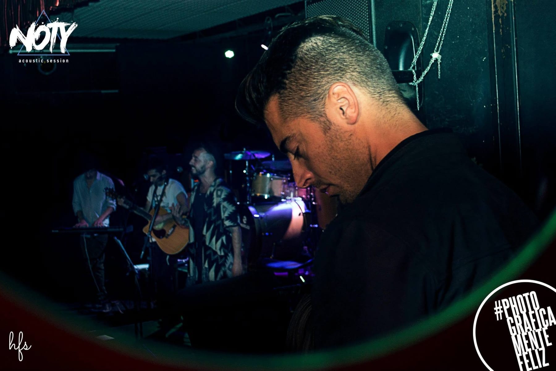 Fotografía a color de un hombre de perfil con pelo corto y remera oscura, cabeza inclinada hacia abajo, con la banda desenfocada tocando detrás bajo una dominante de luz verdosa; cadenas y…