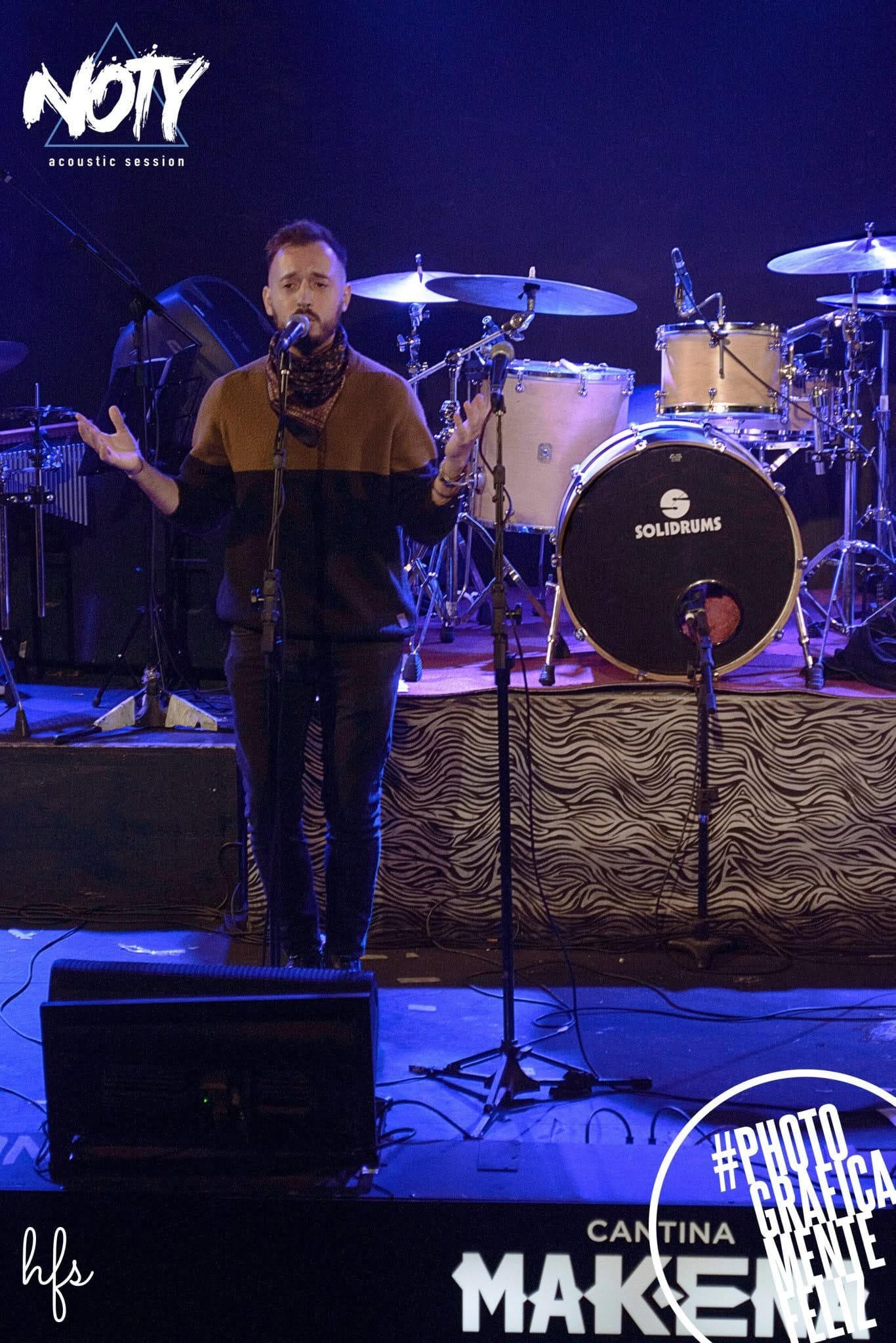 Vista amplia del set de batería y percusión (congas, platillos) sobre el escenario iluminado en azul, con atriles y micrófonos dispuestos, sin músicos visibles en primer plano; instrumentos…
