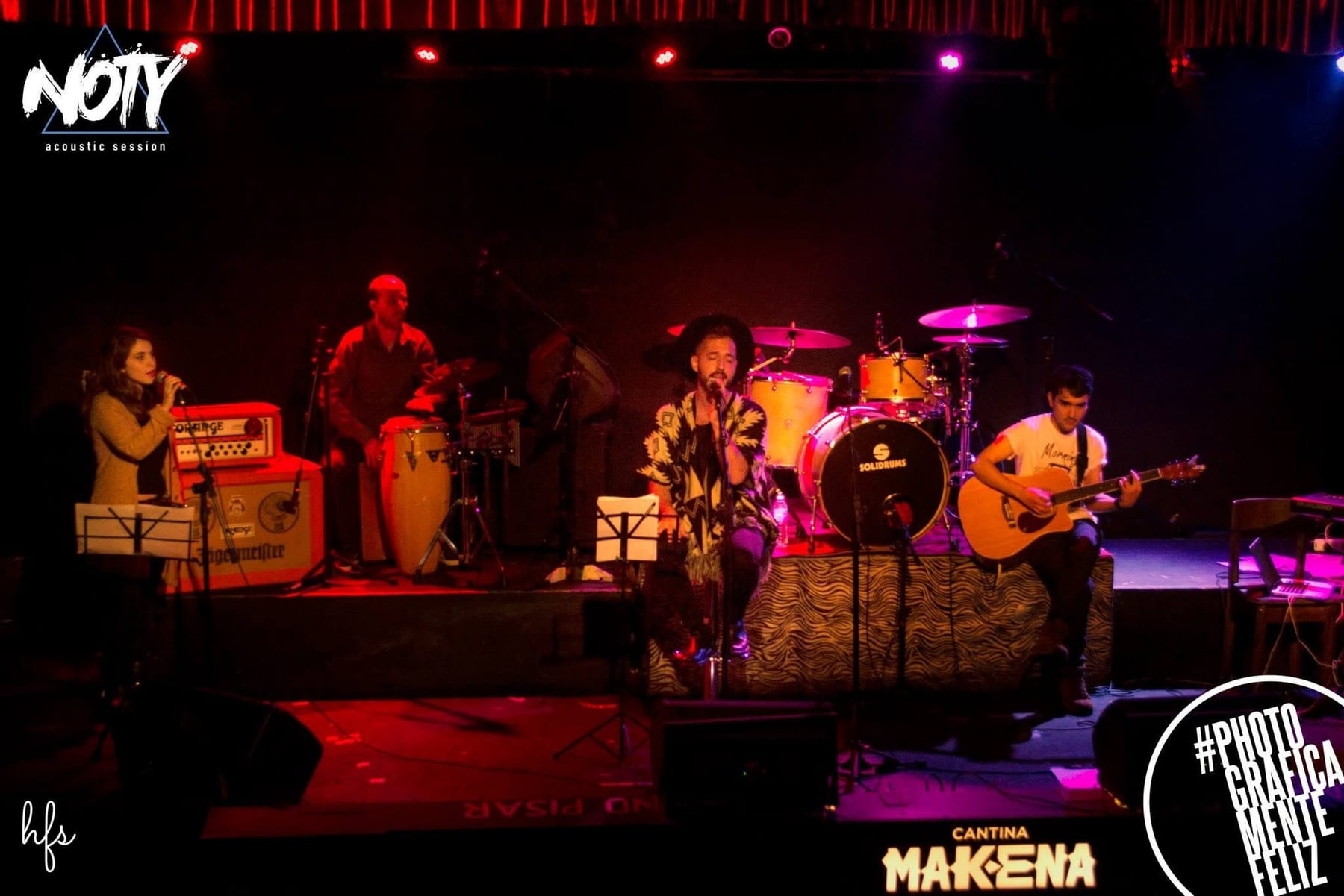 Fotografía a color de la banda completa sobre el escenario: cantante con sombrero, percusionista, baterista, guitarrista y una corista con partitura, bajo un cartel luminoso que dice…