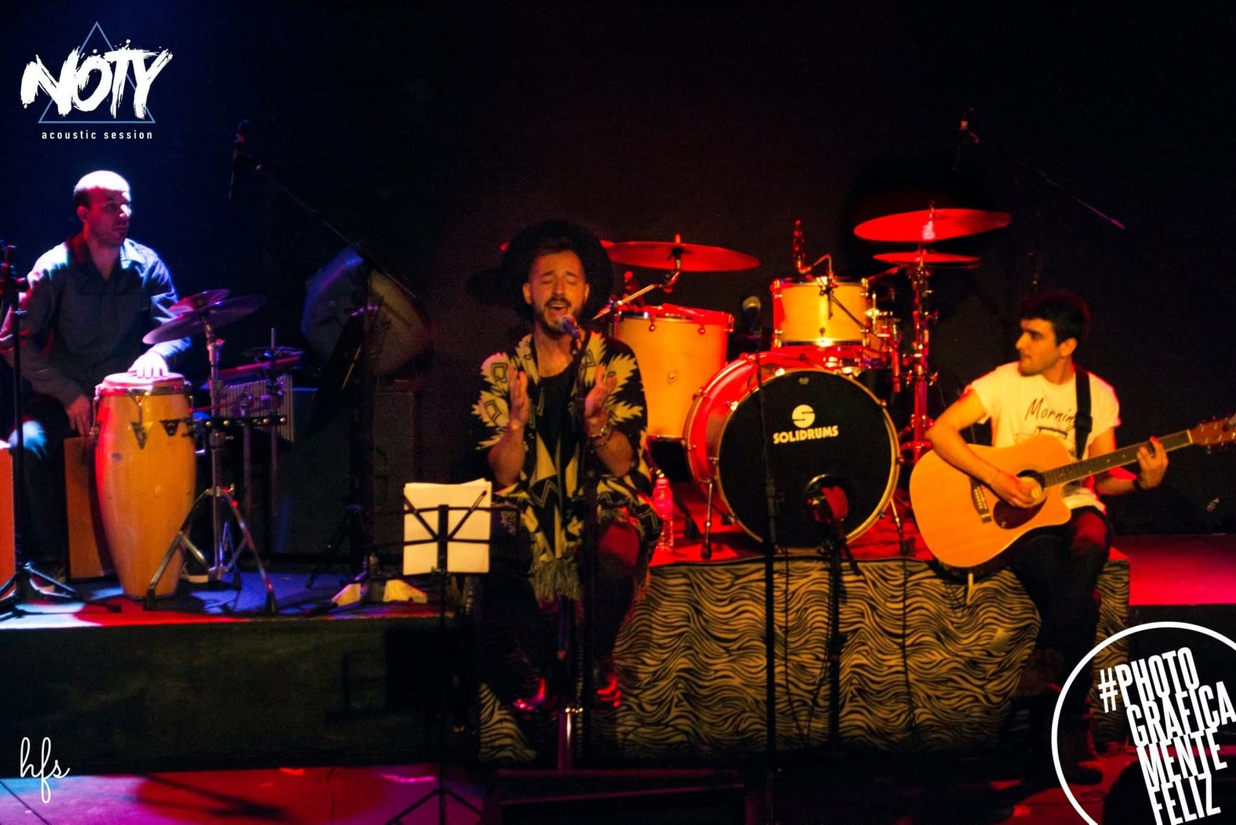 El vocalista canta de frente con un pañuelo estampado al cuello, mientras detrás se distinguen el percusionista y el baterista bajo luces azules.