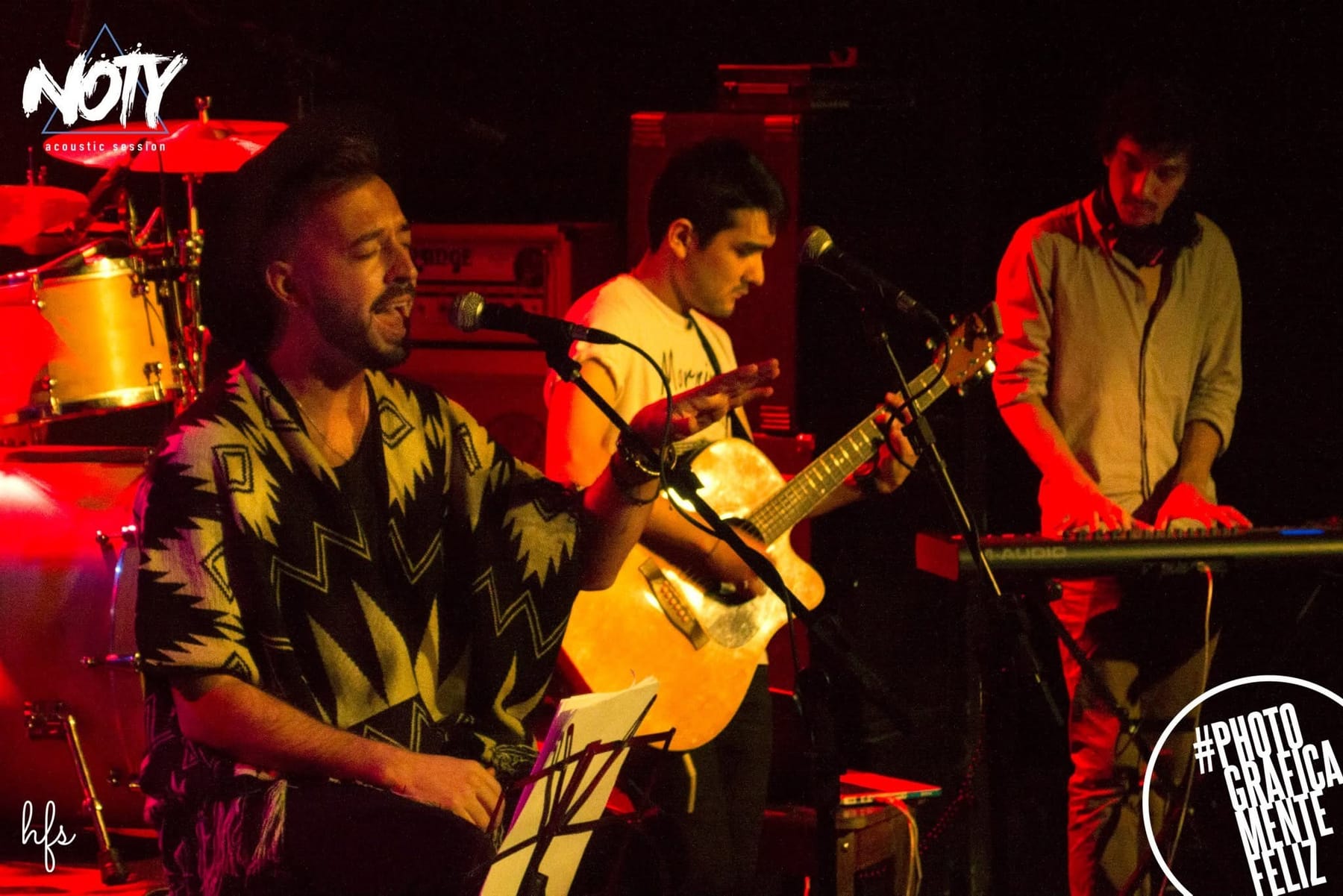 Cantante con poncho estampado sentado en un taburete cantando ante el micrófono, junto a un guitarrista de remera clara tocando guitarra acústica, ambos bañados en luz roja cálida de…
