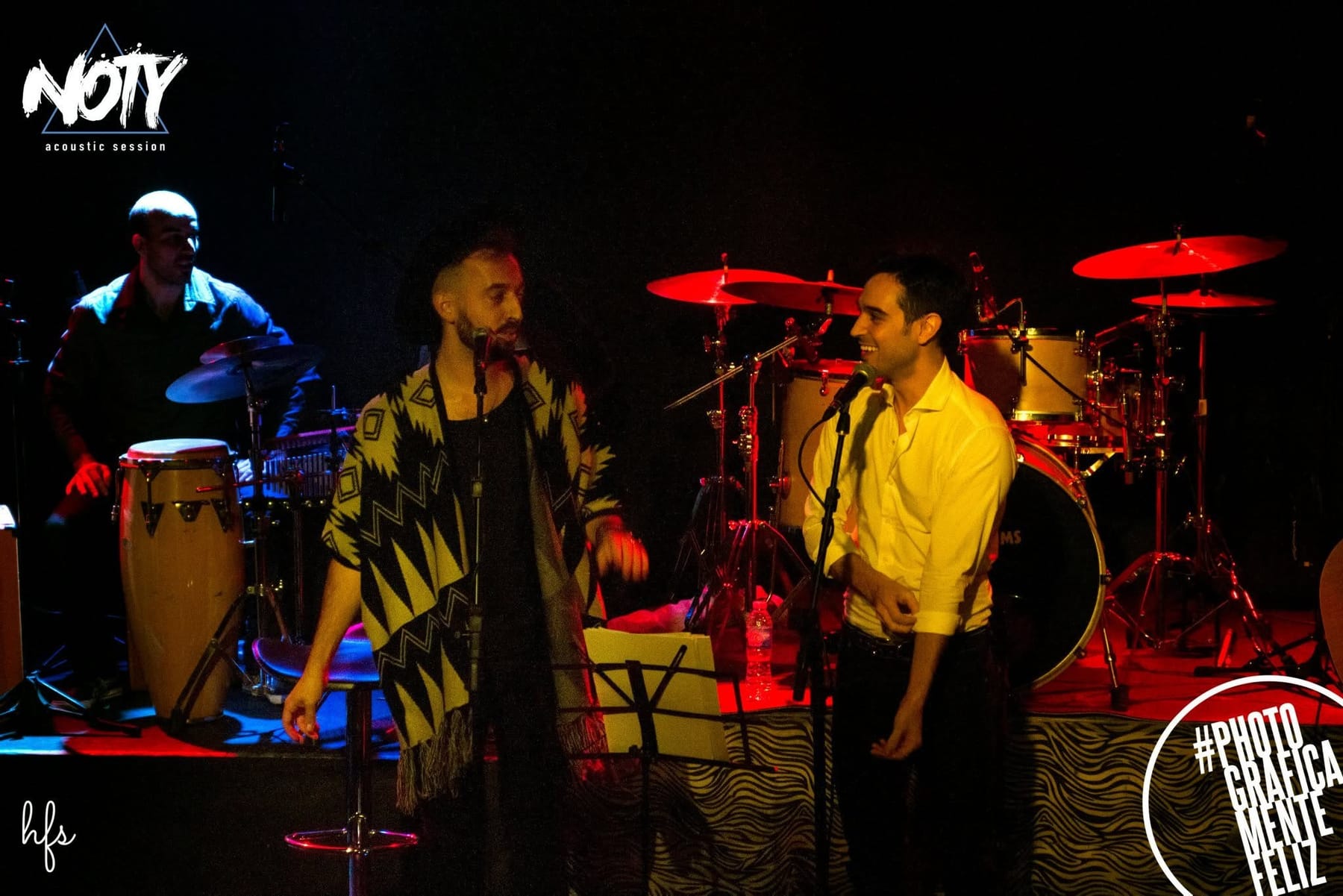 Fotografía a color de dos cantantes conversando de perfil frente a sus micrófonos sobre el escenario, uno con sombrero y poncho y el otro con camisa blanca sonriendo; detrás, un baterista…