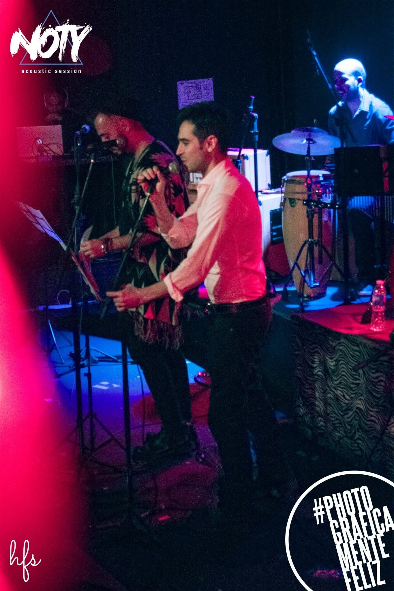 Dos cantantes en el escenario, uno con poncho estampado y otro con camisa blanca, cantan juntos frente a sus micrófonos con expresión concentrada.