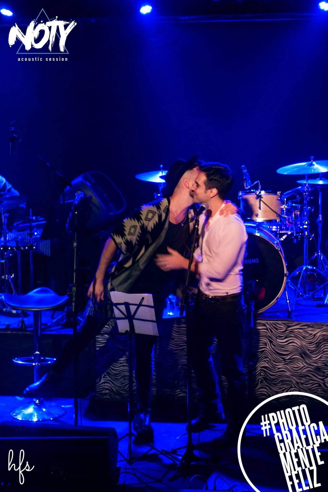 Dos cantantes en el escenario, uno con poncho estampado aplaudiendo con los brazos en alto, el otro de camisa blanca cantando junto al micrófono, bajo un intenso foco de luz blanca y…
