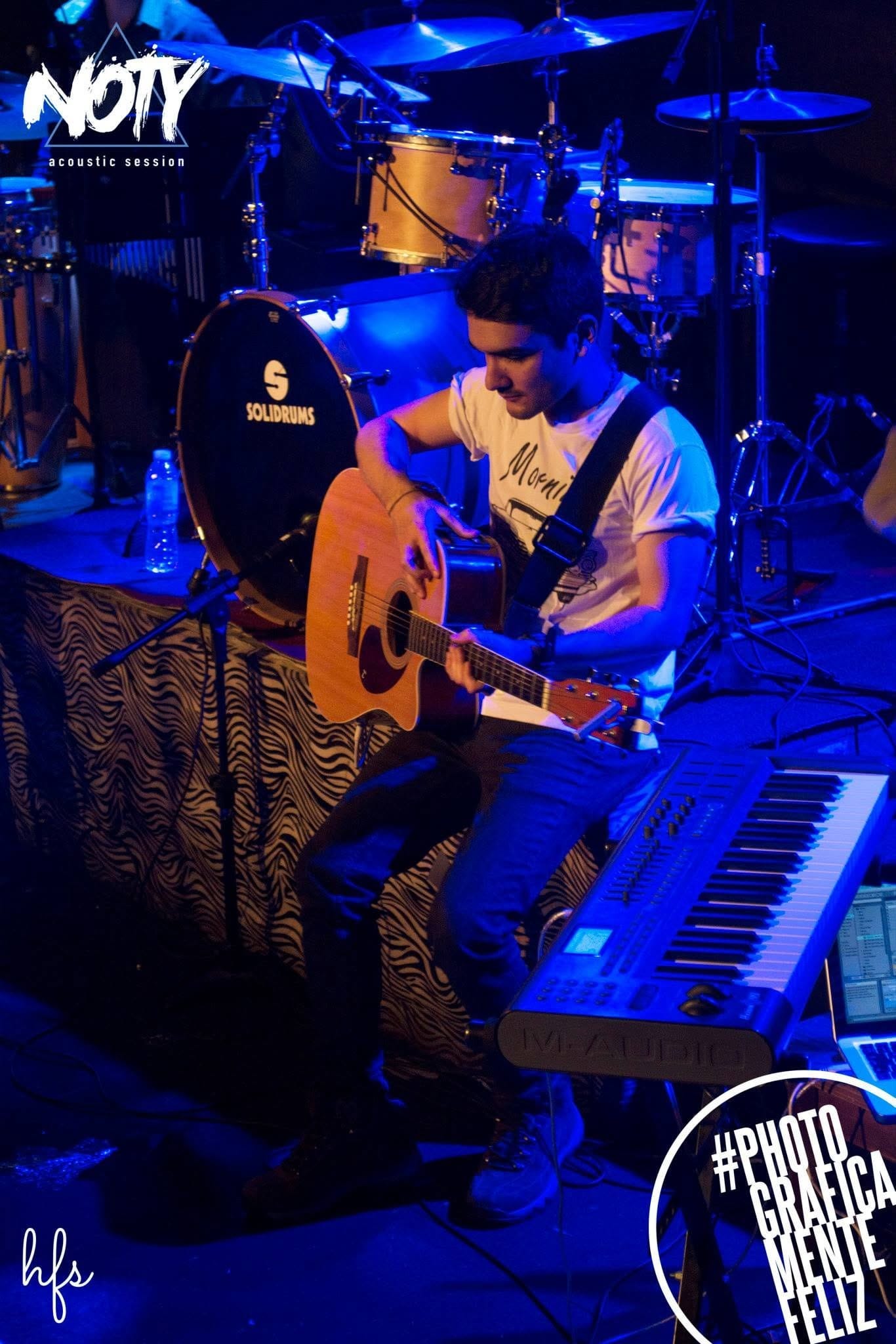Fotografía a color de un guitarrista sentado tocando una guitarra acústica, con remera clara, bajo una fuerte dominante de luz azul; detrás, una batería con parche "SOLIDRUMS" y un teclado…