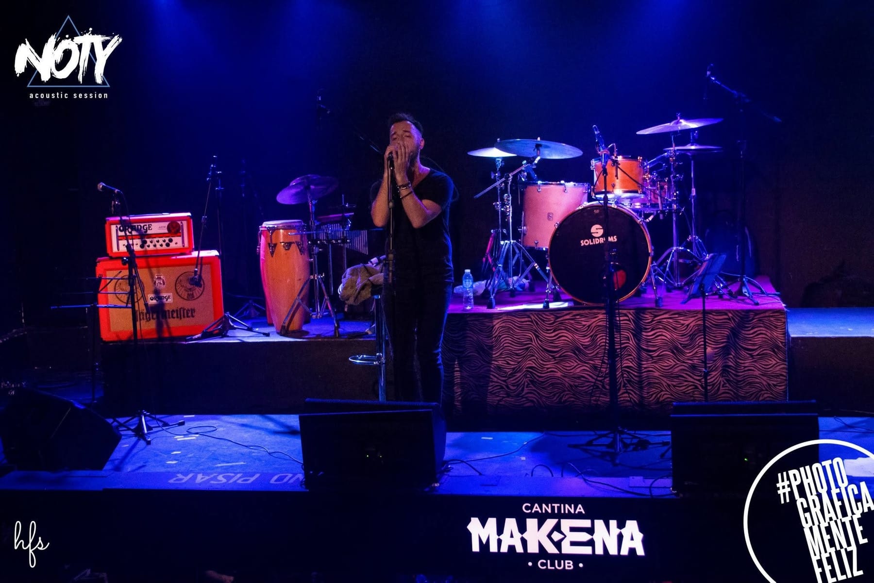 Vista general de un escenario de club nocturno iluminado en azul, con batería "Solidrums", congas, amplificadores Orange y cartel "NOTY acoustic session"; sobre el frente del escenario se…