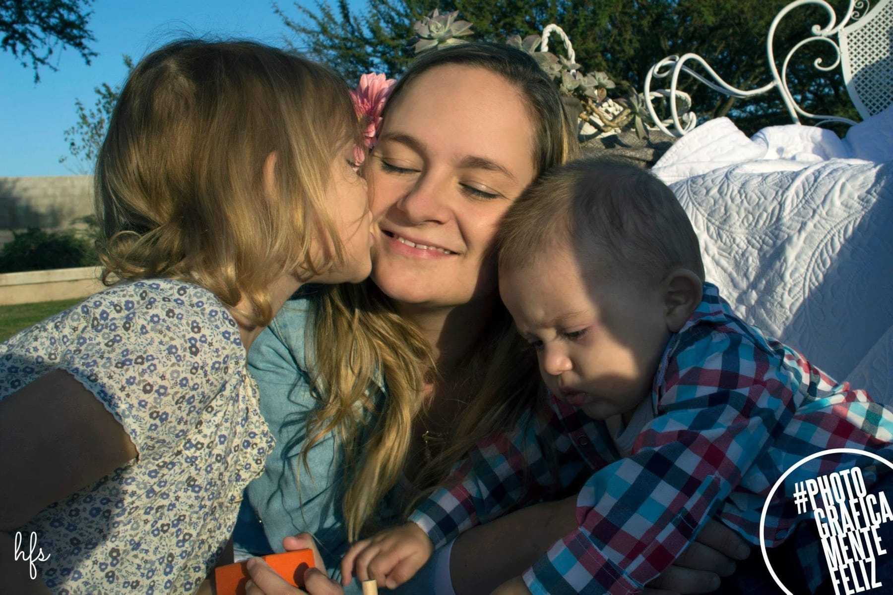 Una madre sonríe con los ojos cerrados mientras recibe un beso de su hija en la mejilla, al tiempo que sostiene en brazos a un bebé que mira hacia abajo.