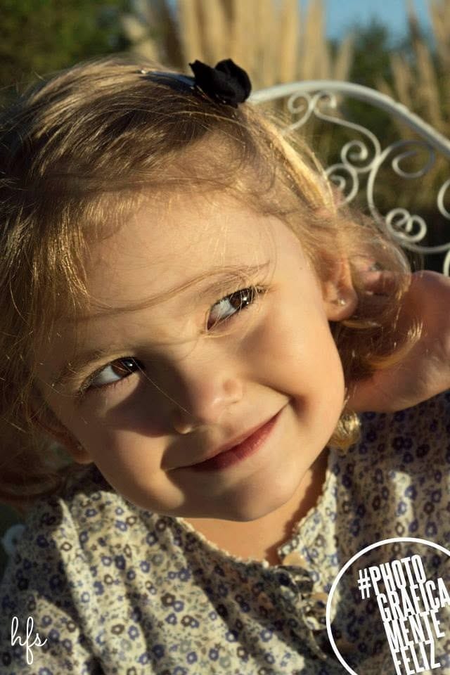 Primer plano de una niña pequeña con rulos rubios y un moño negro en el pelo, mirando de reojo con una sonrisa pícara bajo luz cálida de atardecer.