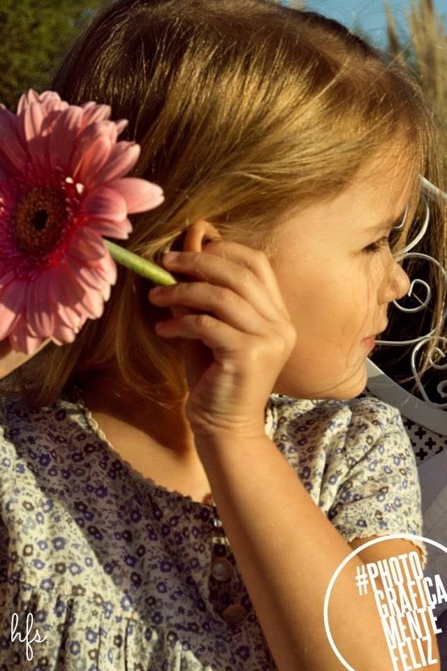 Primer plano de una niña de perfil sosteniendo una gerbera rosa junto a su oreja, con el pelo suelto iluminado por luz de atardecer y vegetación desenfocada al fondo.
