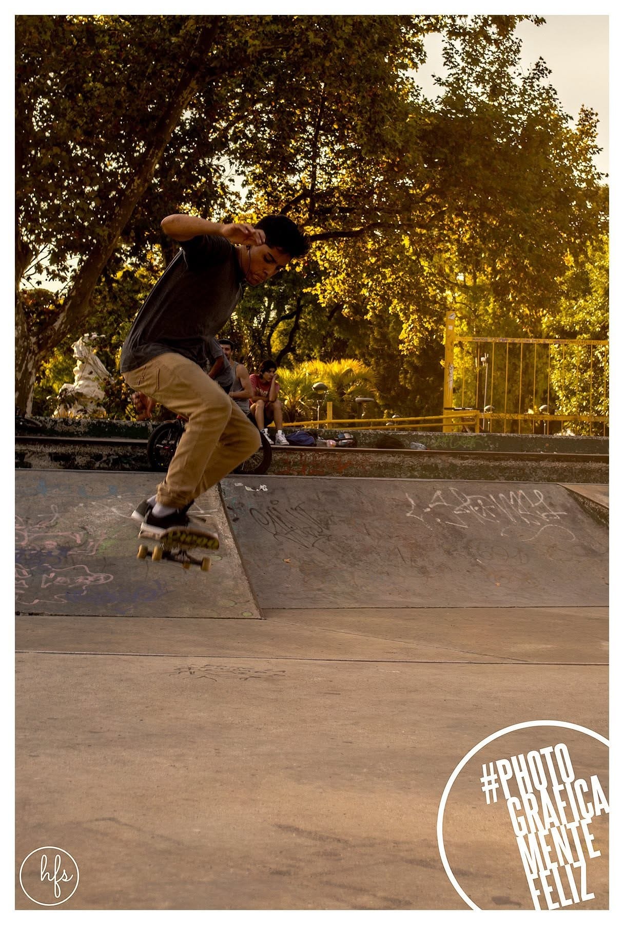 Fotografía a color de un skater saltando desde un borde de cemento grafiteado, con los brazos extendidos en pleno movimiento; al fondo, árboles iluminados por luz de atardecer y otras…