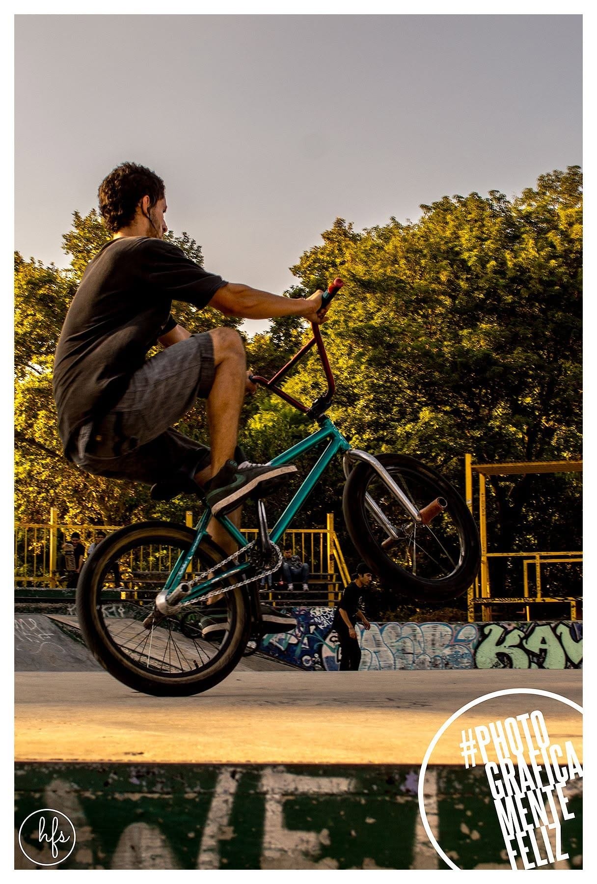 Ciclista de BMX en wheelie sobre una rampa de skatepark, remera oscura y short, rodeado de árboles dorados por la luz de atardecer; graffiti visible en la base de la rampa.