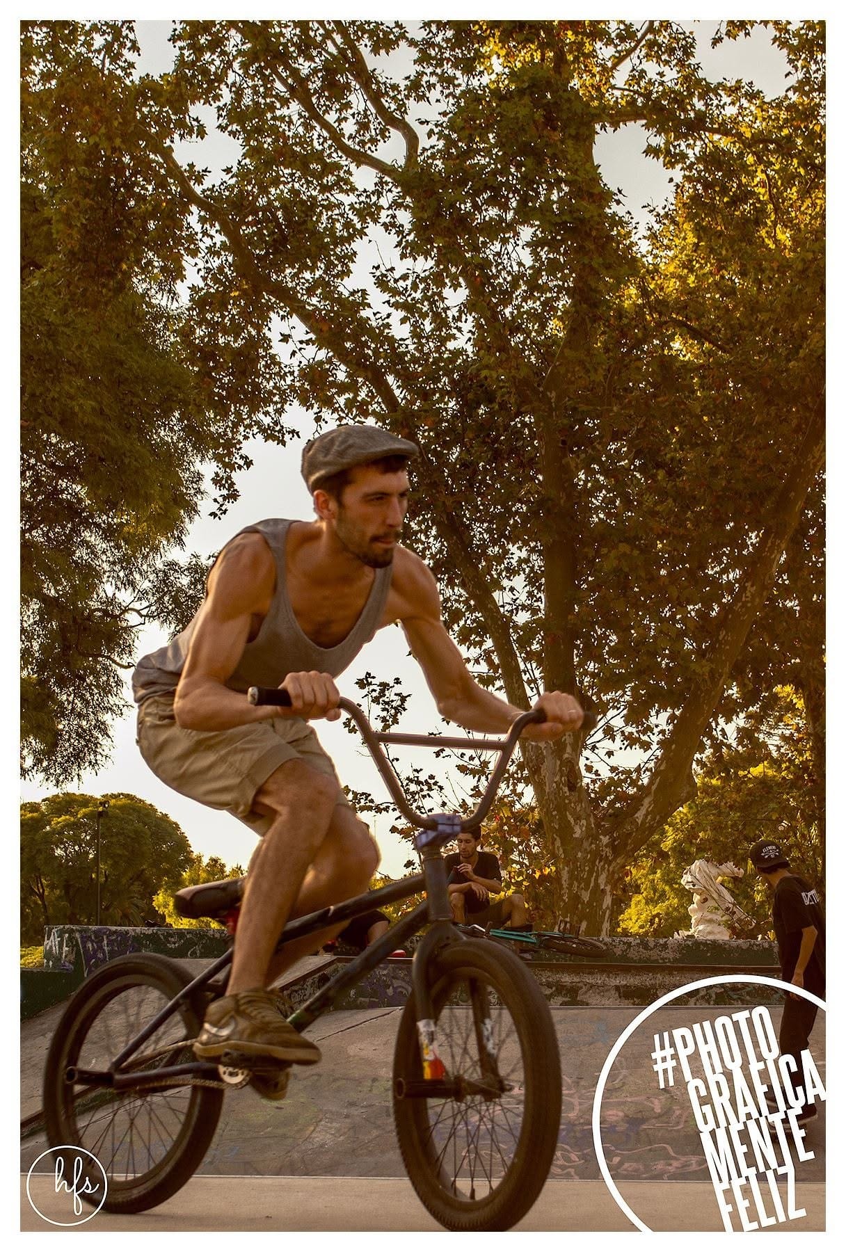 Ciclista de BMX con boina y musculosa gris, de pie sobre los pedales en plena rampa, con la luz del atardecer atravesando el follaje detrás.