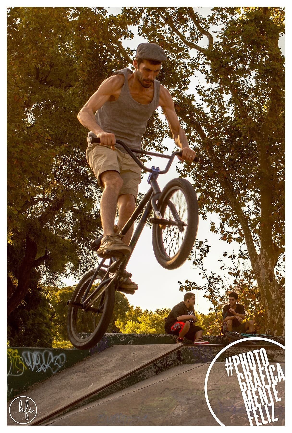 Ciclista de BMX en pleno salto sobre una rampa, con gorra tipo boina, musculosa gris y short beige, brazos tensos sobre el manubrio, fotografiado en contrapicado con árboles y dos…