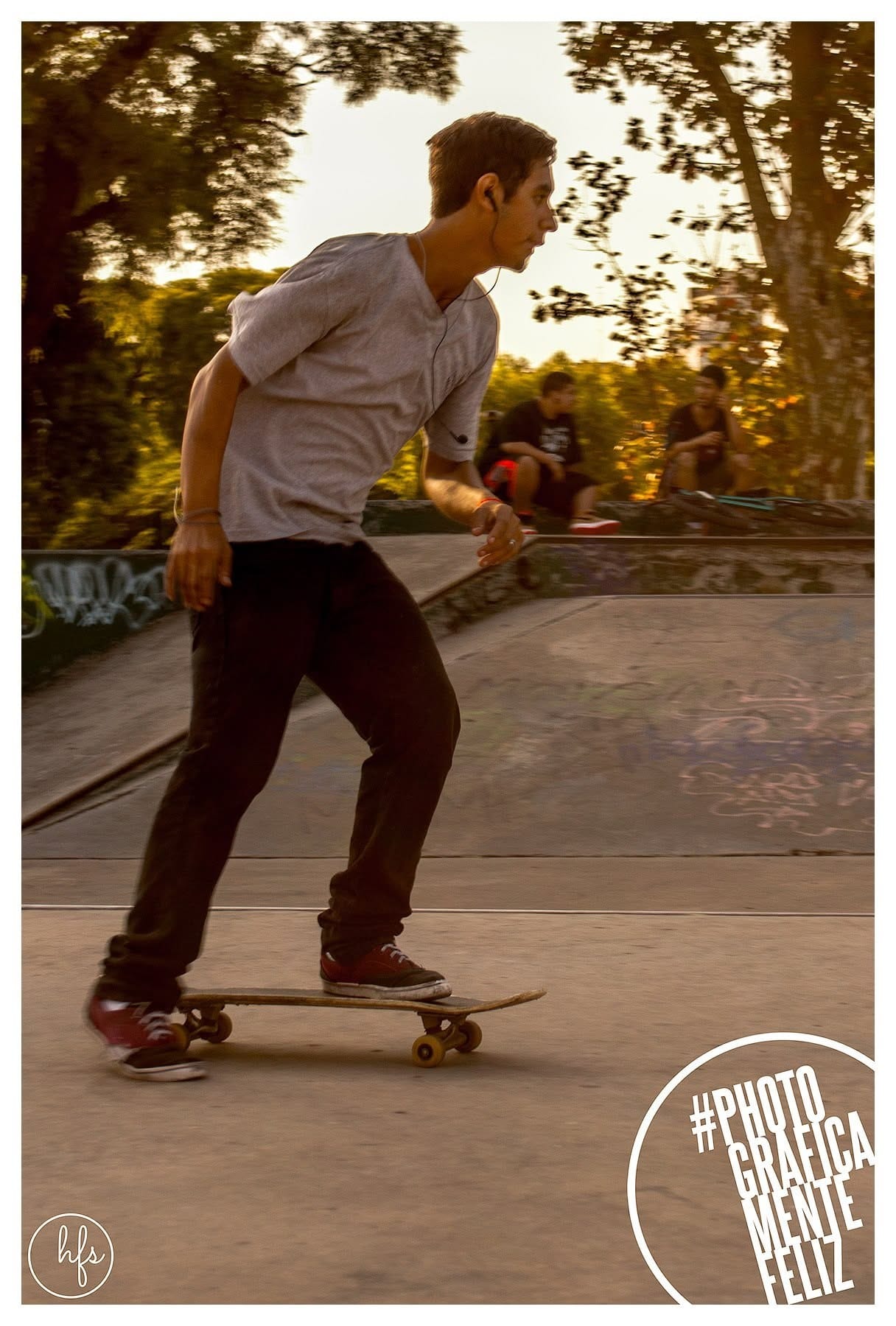 Hombre joven en perfil andando en skateboard sobre pavimento, con auriculares puestos, remera gris y jean oscuro, iluminado por la luz dorada del atardecer que recorta su figura contra…