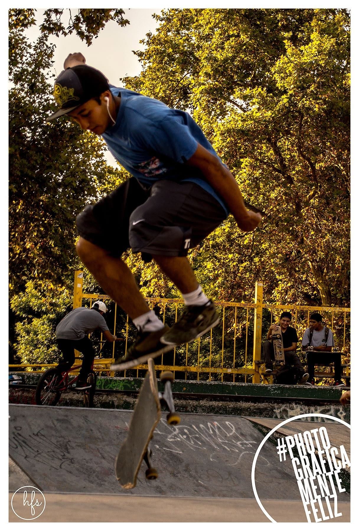Skater en pleno salto sobre una rampa de un skatepark, con gorra hacia atrás, remera azul y auriculares, tabla capturada en el aire bajo árboles iluminados por luz dorada de atardecer…