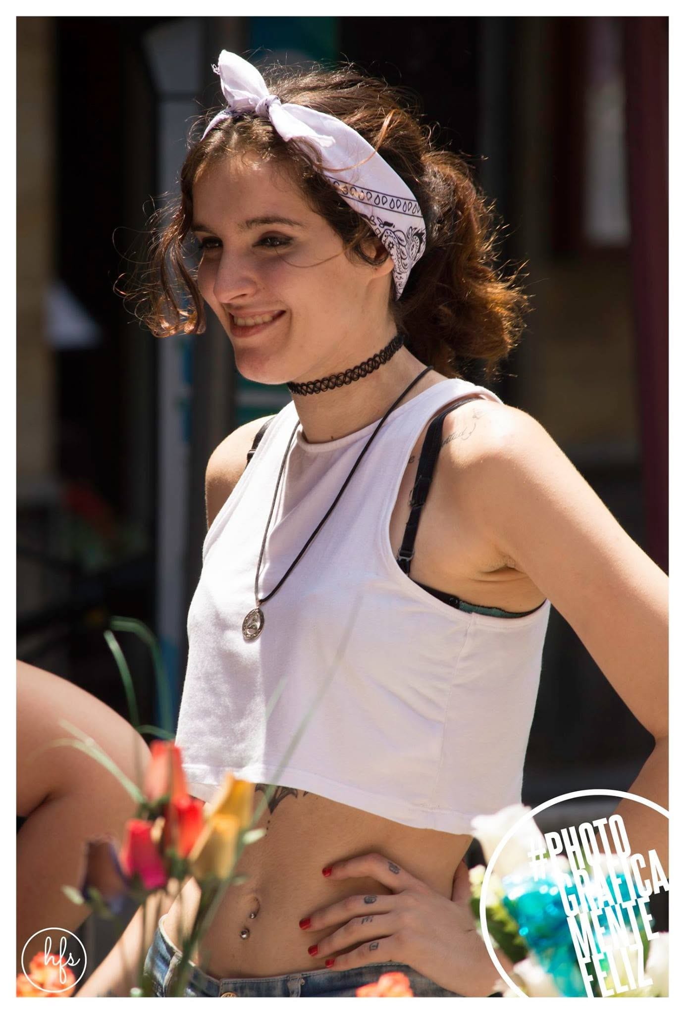 Retrato a color de una joven con pañuelo blanco anudado en la cabeza, gargantilla negra tipo tatuaje y top blanco corto, con tatuajes visibles en el vientre; sostiene ramas de rosas en…