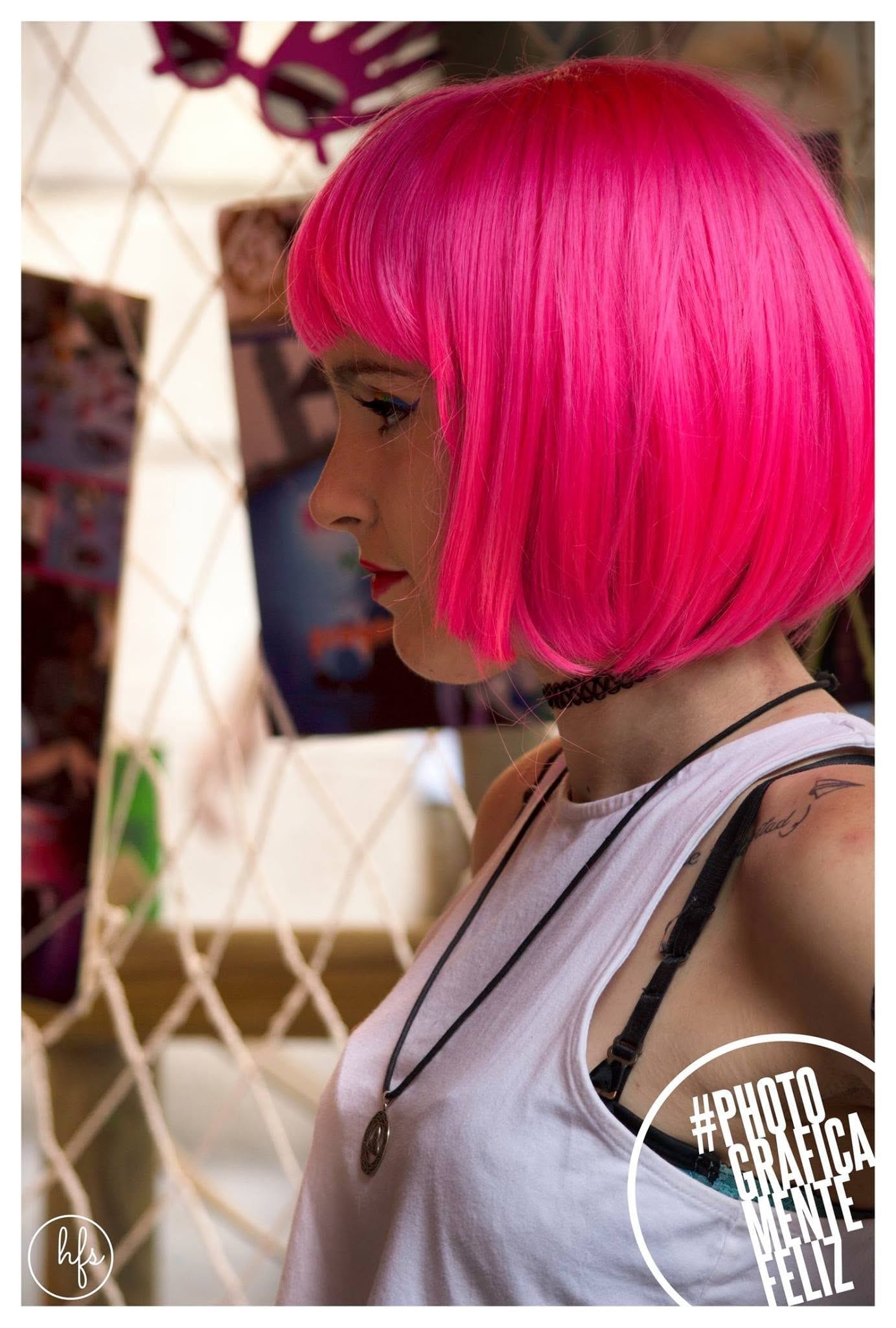 Retrato de perfil de una persona con peluca bob rosa flúo, labios rojos, gargantilla y musculosa blanca, frente a un fondo con red decorativa y fotografías colgadas.