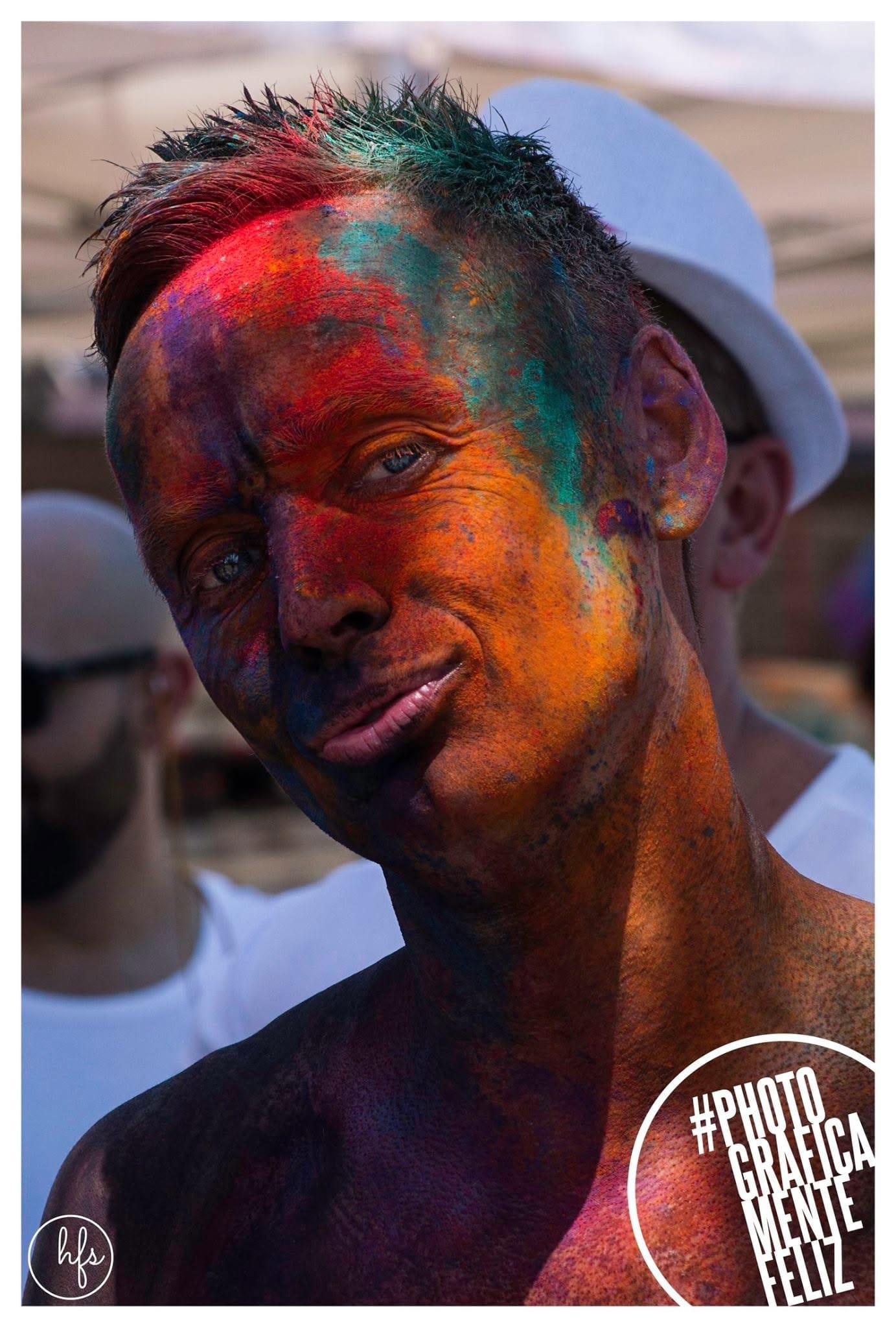 Retrato de un hombre con el rostro y el pelo cubiertos de pigmentos rojo, naranja y verde, sonriendo levemente a cámara, gorra blanca de otra persona visible detrás desenfocada.