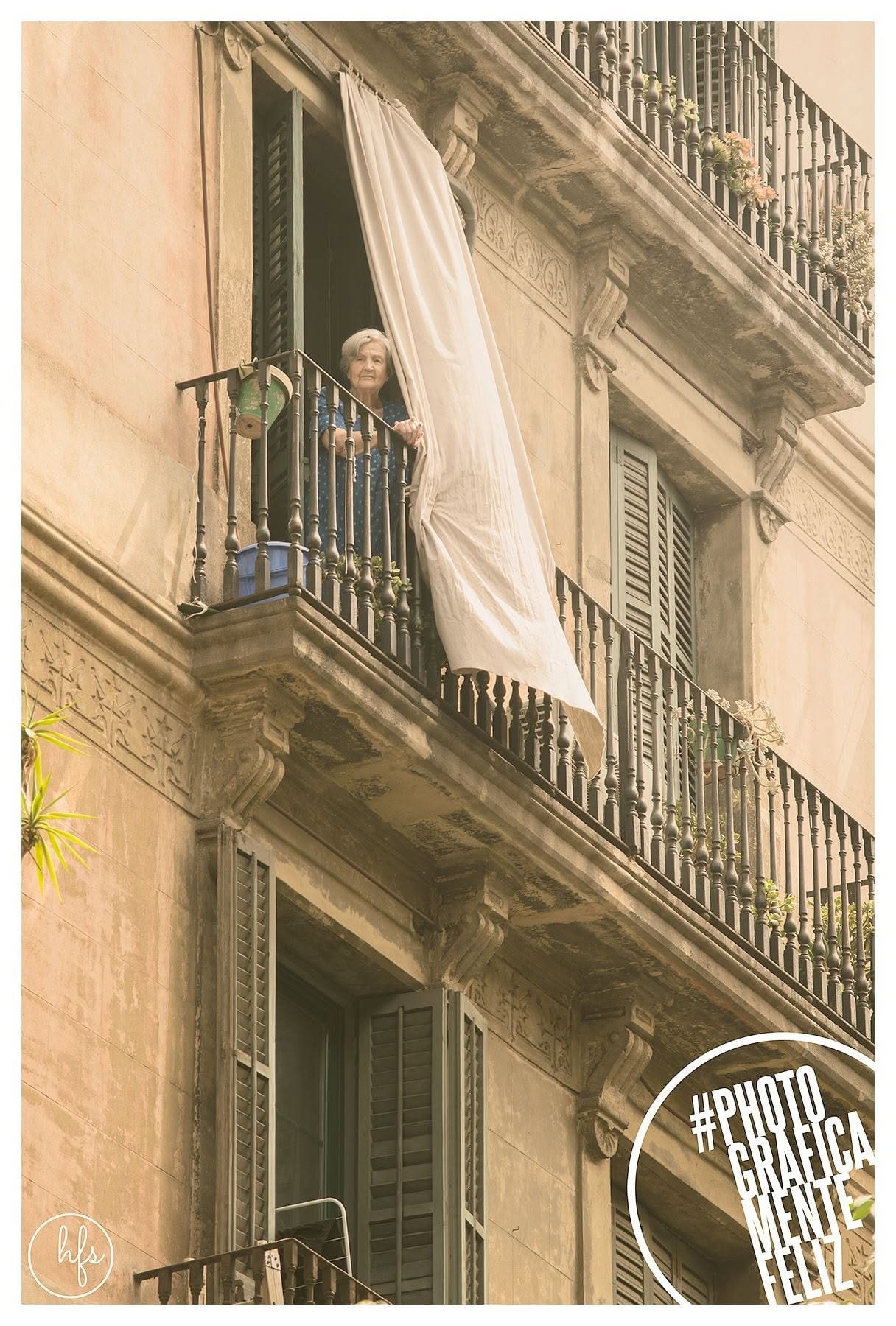 Mujer mayor de pelo blanco asomada a un balcón con una cortina clara ondeando al viento, apoyada en la baranda de hierro de un edificio histórico con fachada ornamentada, en tono sepia…